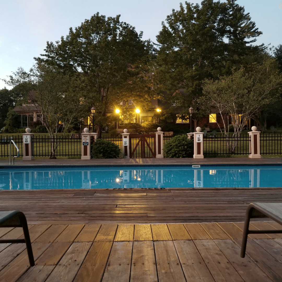 A still swimming pool reflects the evening sky, surrounded by trees and a well-lit house in the background.
