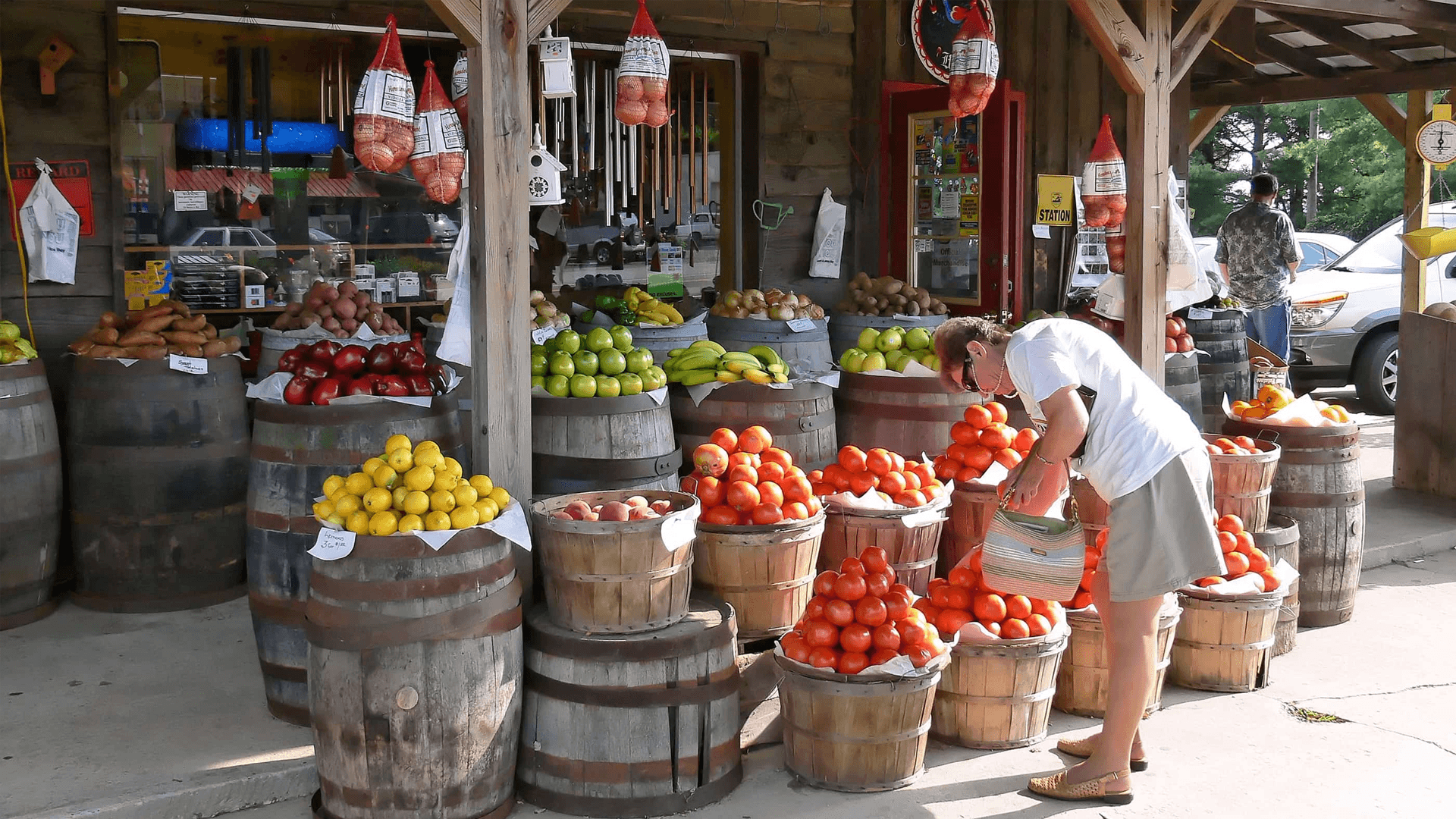 A person selects tomatoes from wooden barrels at an outdoor fruit stand. Vibrant produce includes lemons, apples, and limes, creating a lively, market atmosphere.