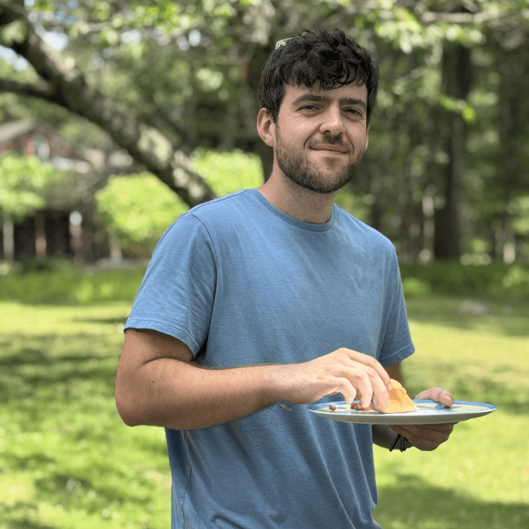 A young man in a blue shirt holds a plate of food while standing in a grassy outdoor setting.