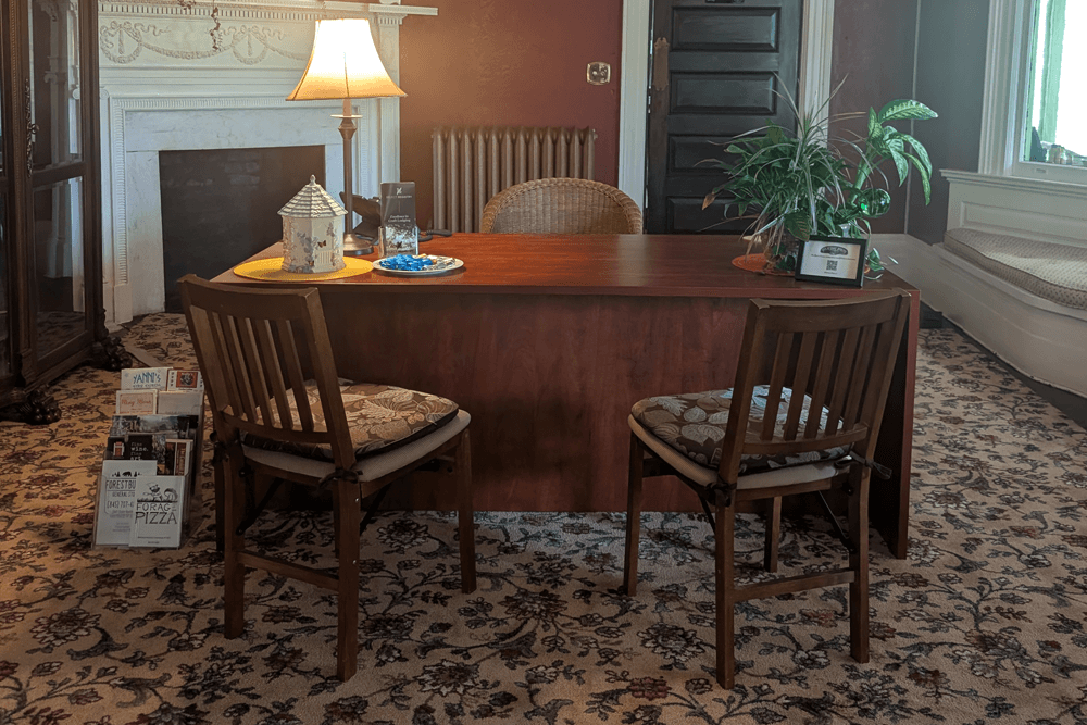 Elegant office with a vintage vibe featuring a wooden desk, two chairs, a chandelier, a lamp, and a plant. Warm tones evoke a cozy, classic feel.