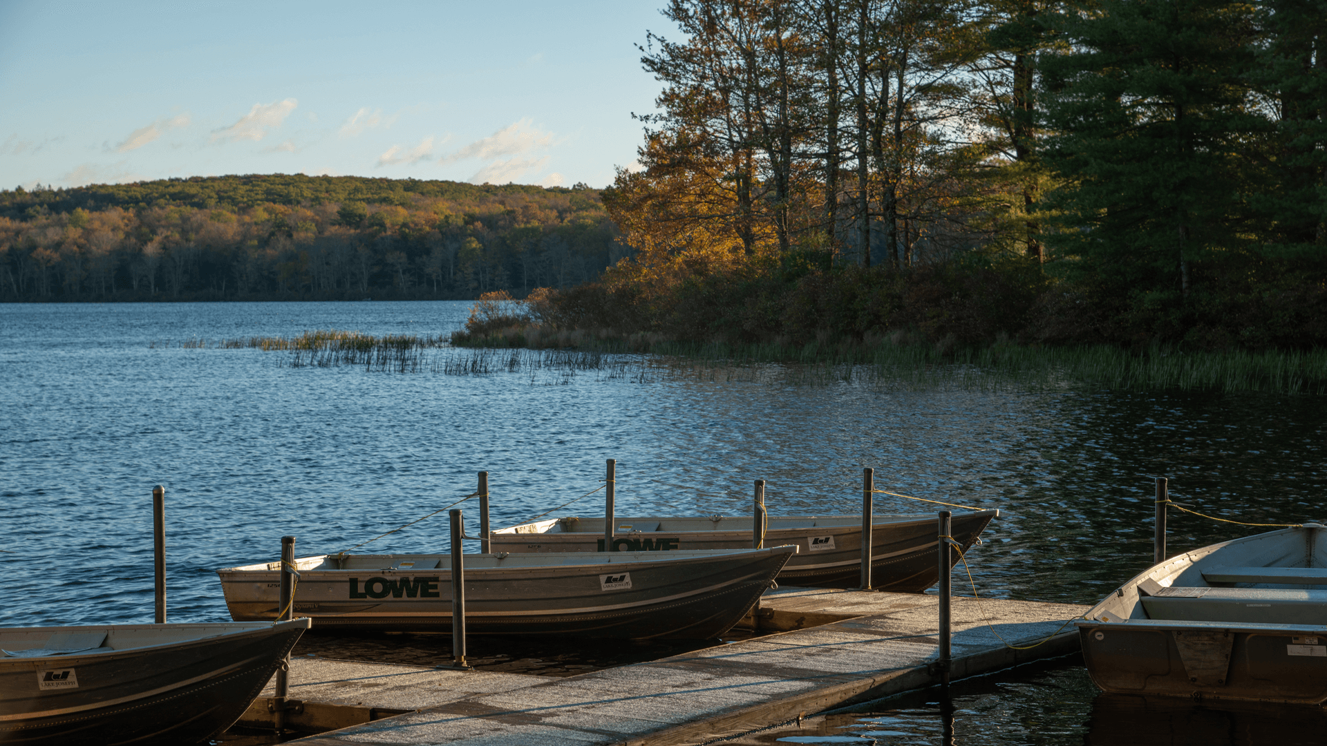 A serene lakeside scene features small boats moored to a wooden dock. Calm water reflects autumn trees under a clear sky, evoking a tranquil mood.
