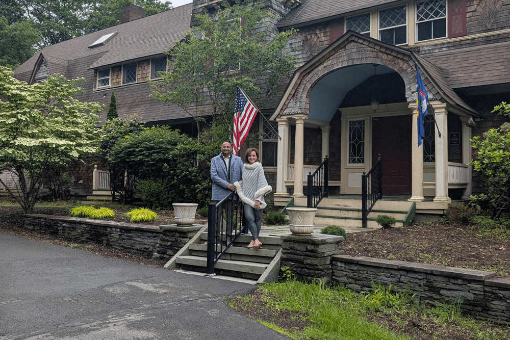 A couple stands together on the steps of a large house with an American flag and greenery surrounding them.