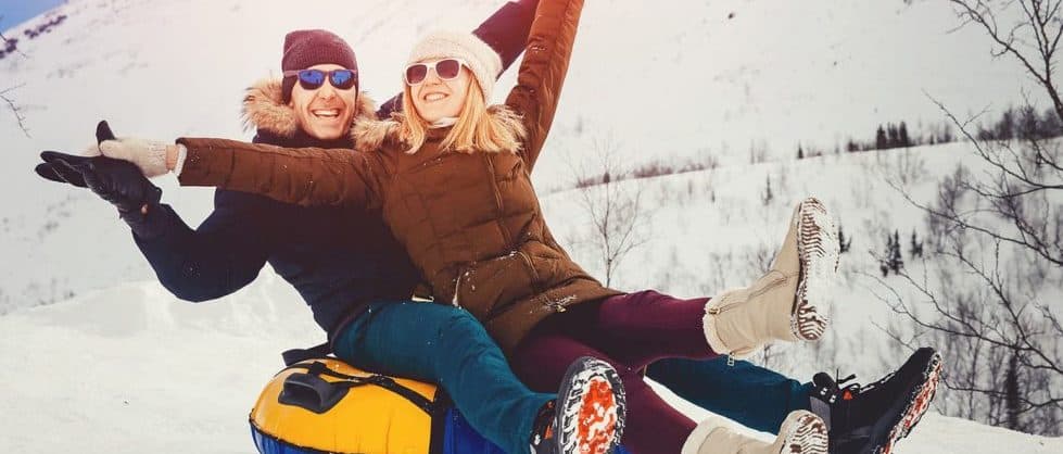 A joyful couple smiles and raises their arms while tubing down a snowy hill.