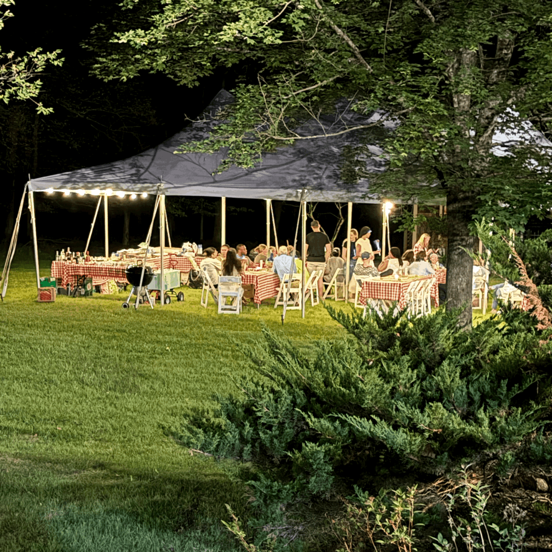 A large outdoor tent is filled with people seated at red-checkered tables, enjoying a night gathering under bright lights.