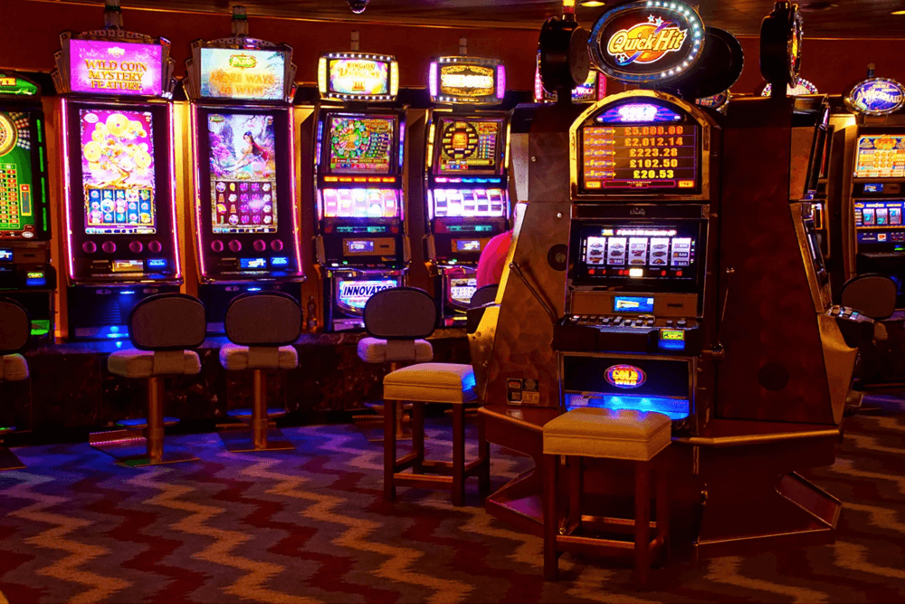 A brightly lit casino area featuring multiple slot machines and empty chairs.