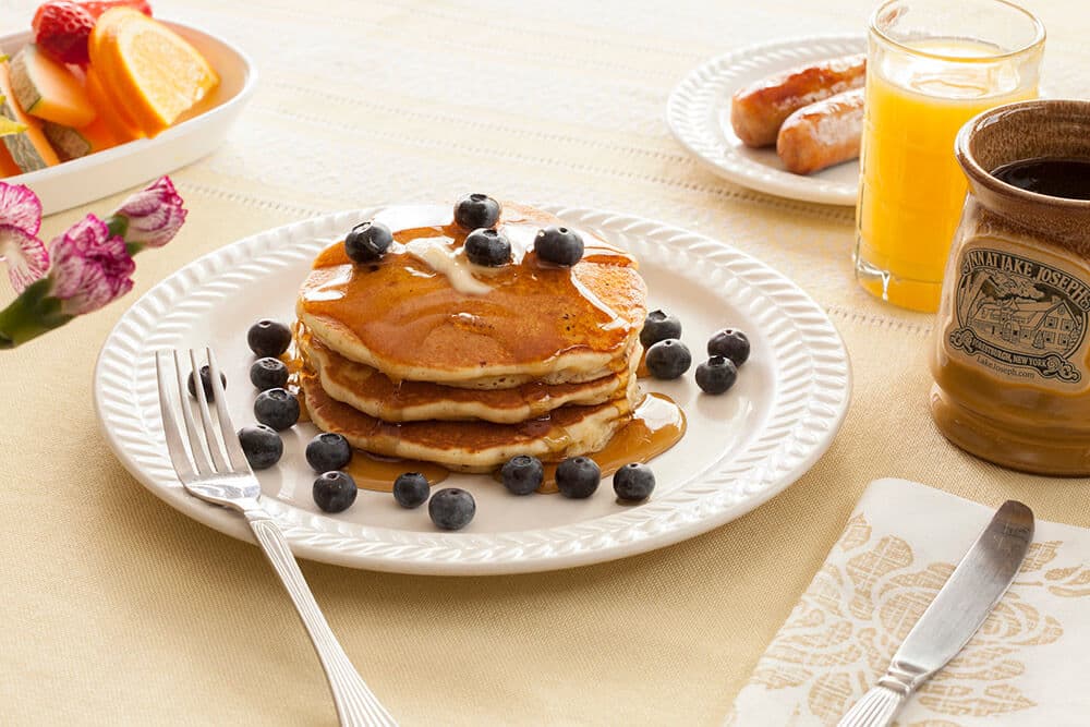 A stack of pancakes topped with butter and blueberries drizzled with syrup sits on a white plate. Nearby are sausages, orange juice, coffee, and fruit slices.