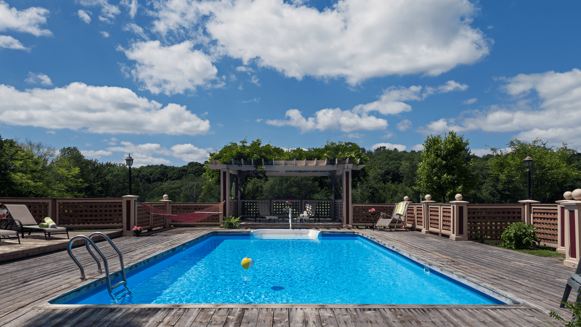 A clear blue swimming pool surrounded by a wooden deck and green trees under a partly cloudy sky.