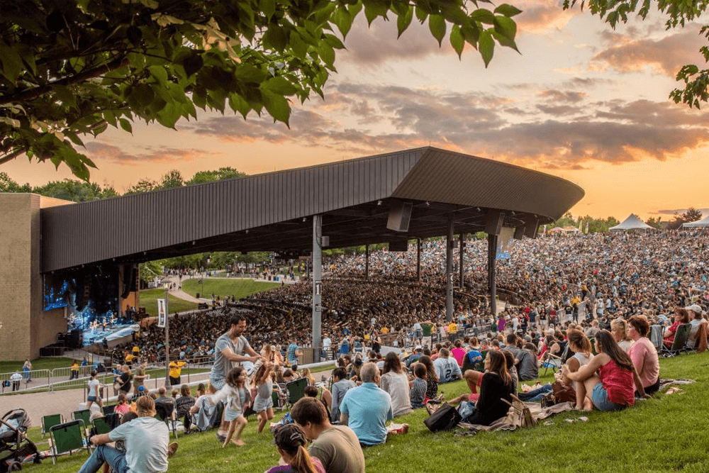 Outdoor concert at sunset with a large crowd seated on a grassy hill. A stage is partially visible under a pavilion. The atmosphere is lively and relaxed.