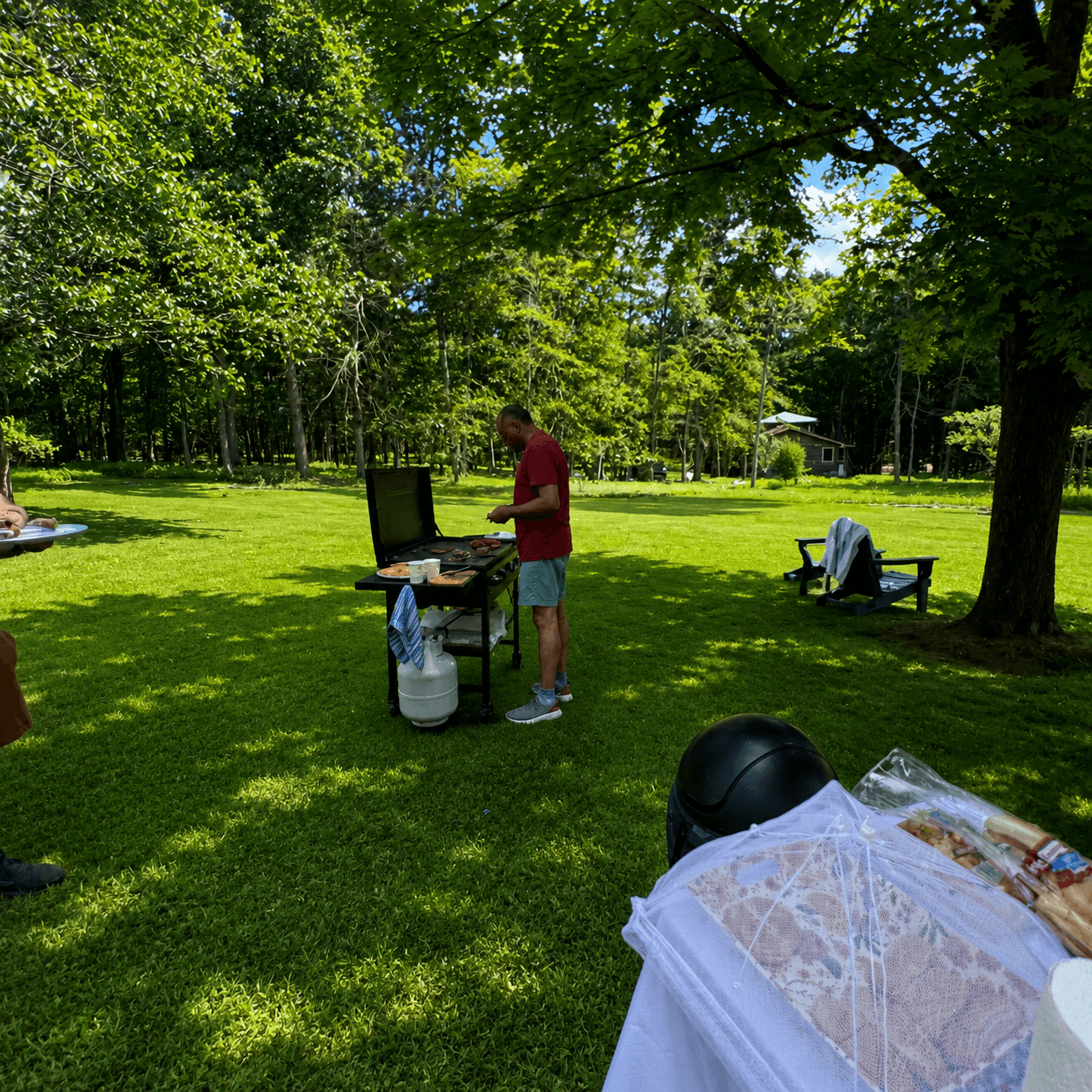A man grills food outdoors in a grassy area surrounded by trees.