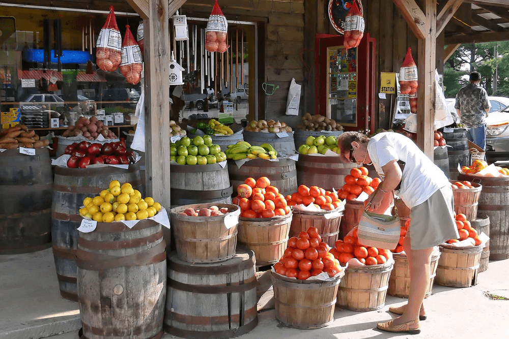 A woman in a white shirt and mask sorts through baskets of colorful fruits at a market stall.