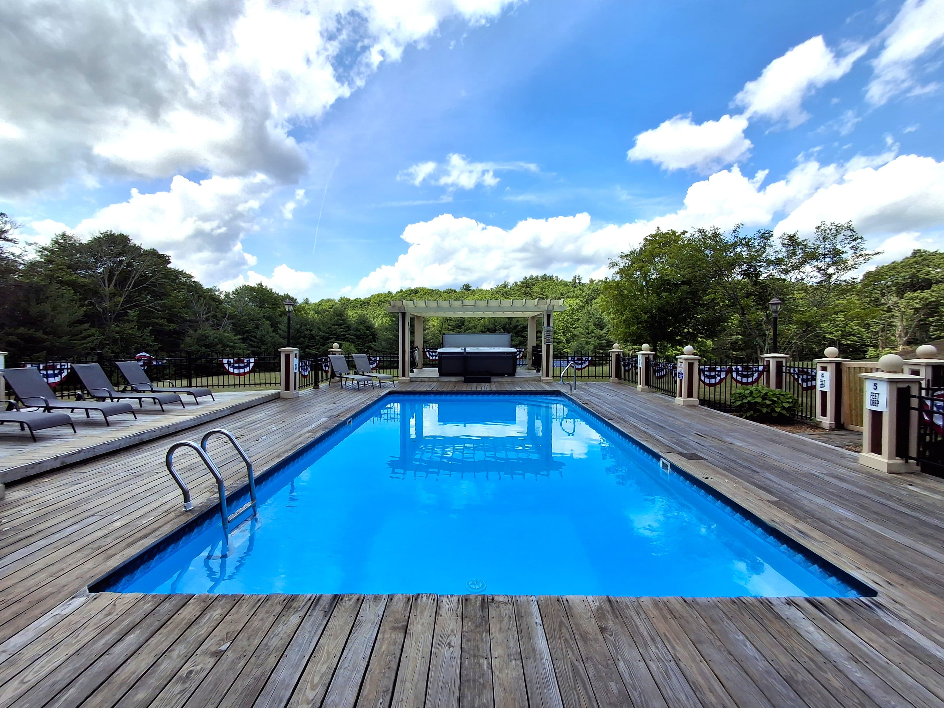 A bright blue pool surrounded by wooden decking and lounge chairs, set against a backdrop of green trees and a cloudy sky.
