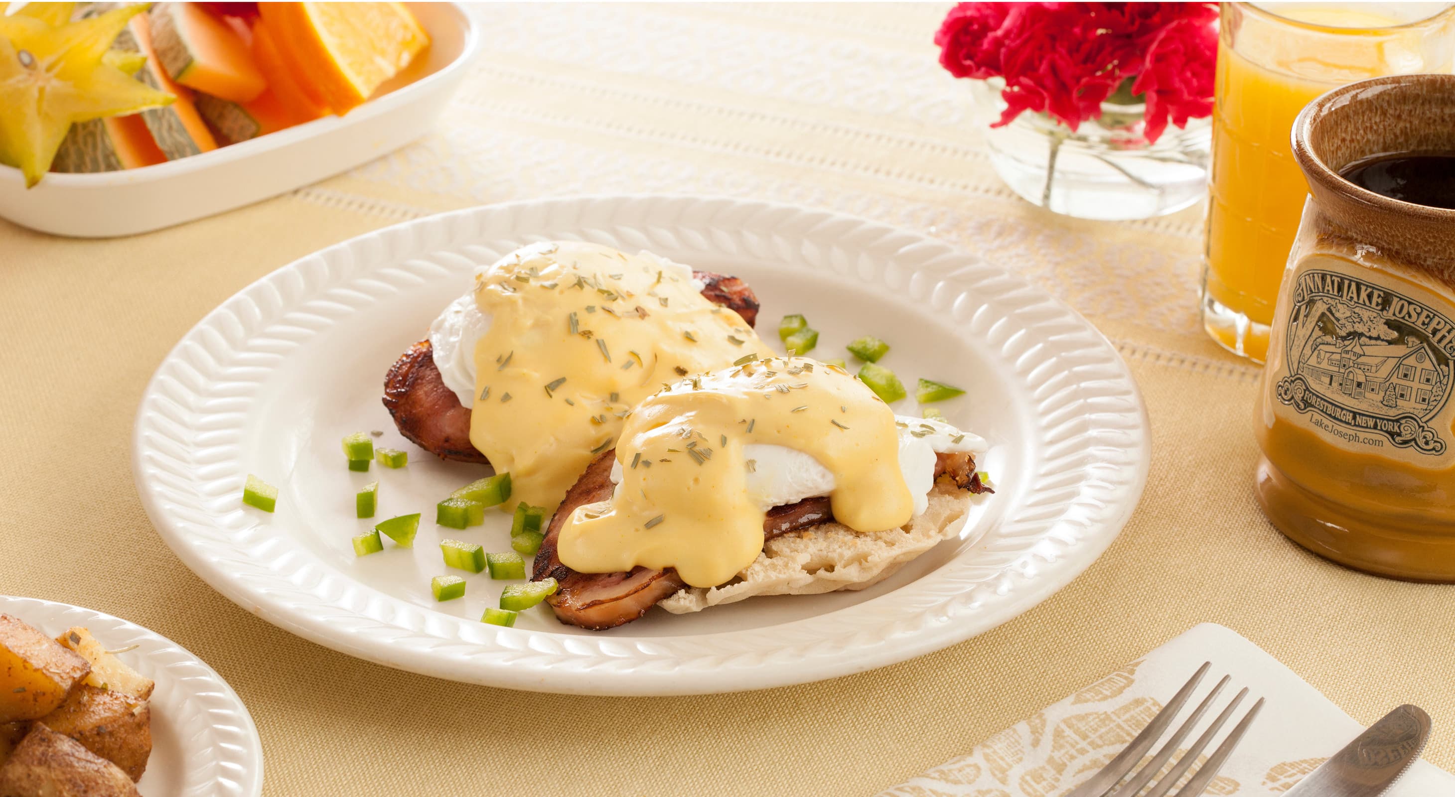 Plate with eggs Benedict topped with creamy hollandaise sauce, garnished with herbs. Accompanied by a fruit bowl, coffee, and orange juice on a beige tablecloth.