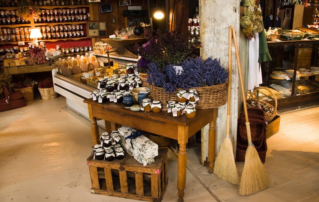 A rustic market display features jars of preserves, floral arrangements, and handmade goods on a wooden table beside baskets and cleaning brooms.