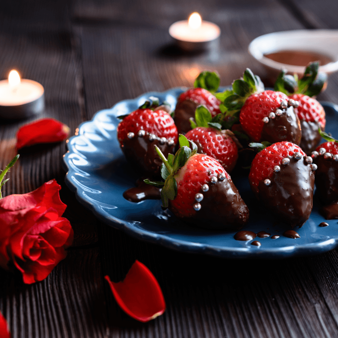 A plate of chocolate-covered strawberries adorned with silver sprinkles, surrounded by rose petals and candles.