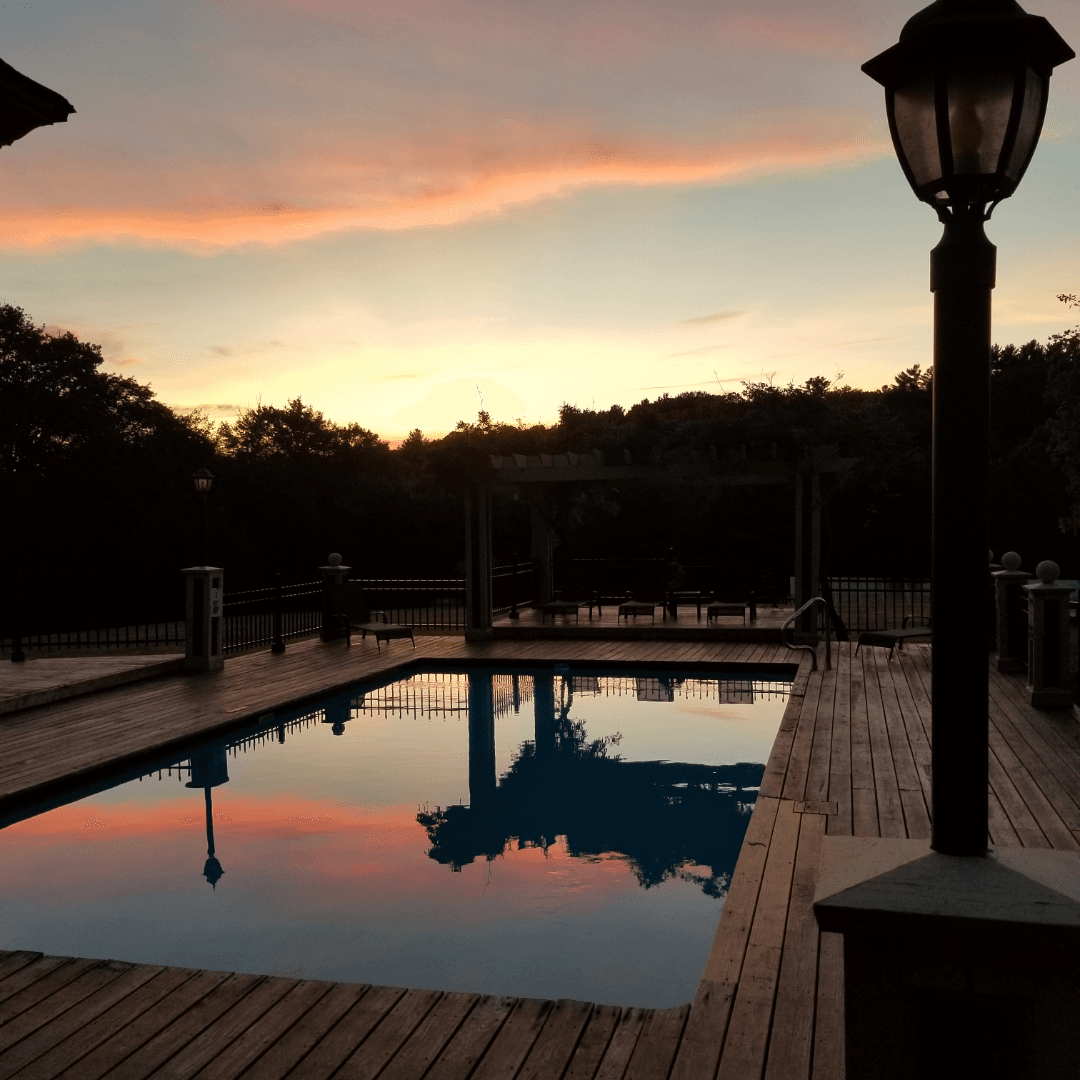 A serene outdoor pool reflects the colorful sunset sky, surrounded by wooden decking and greenery.