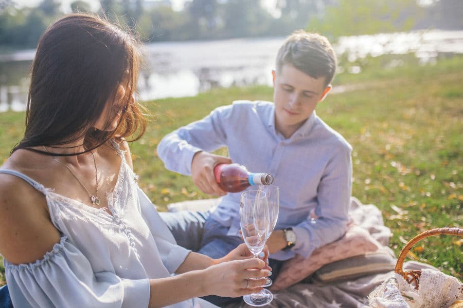 A young couple enjoys a picnic by the water, with one pouring rosé wine into a glass.