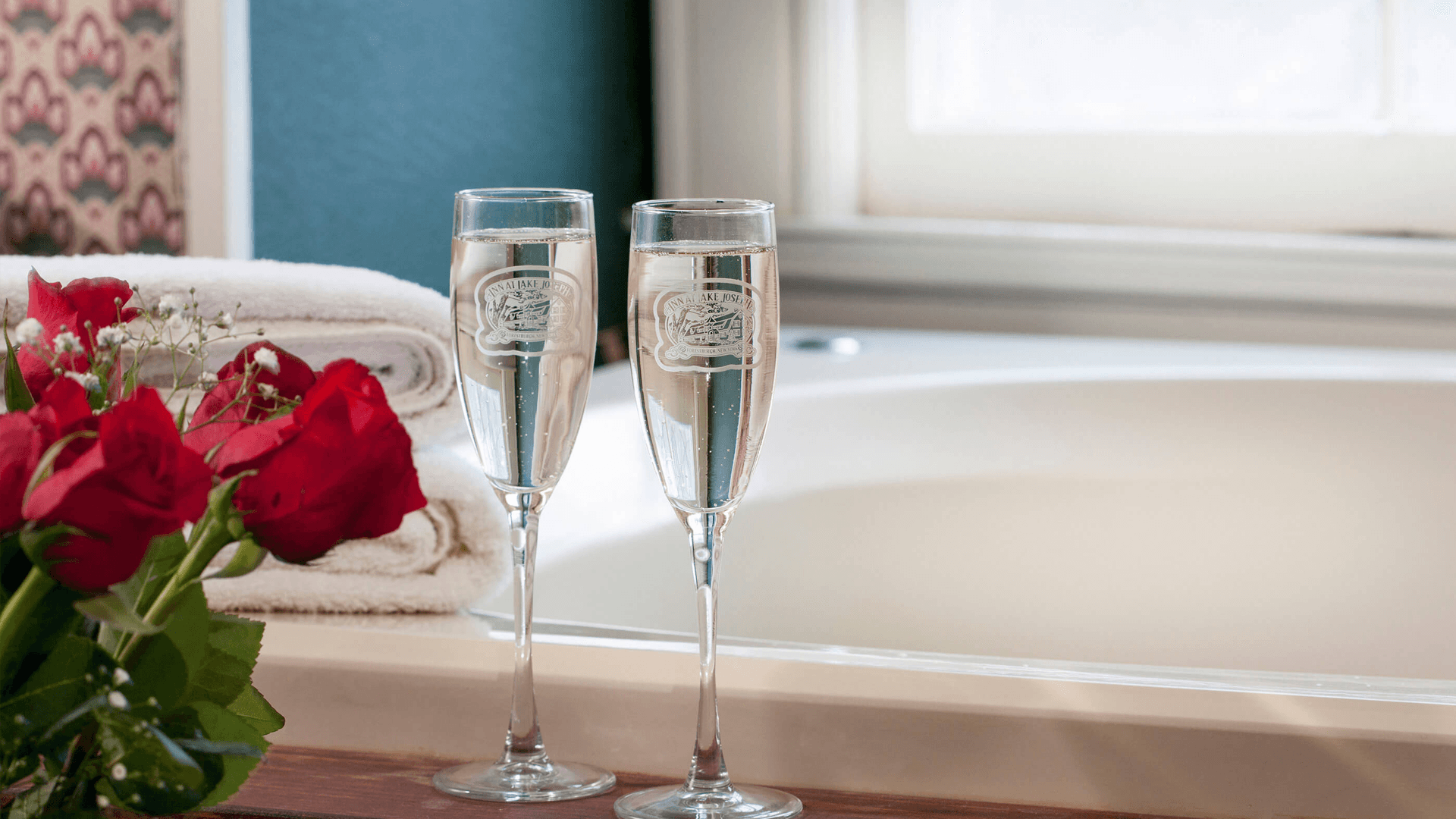 Two champagne flutes beside a bouquet of red roses and rolled towels near a bathtub.