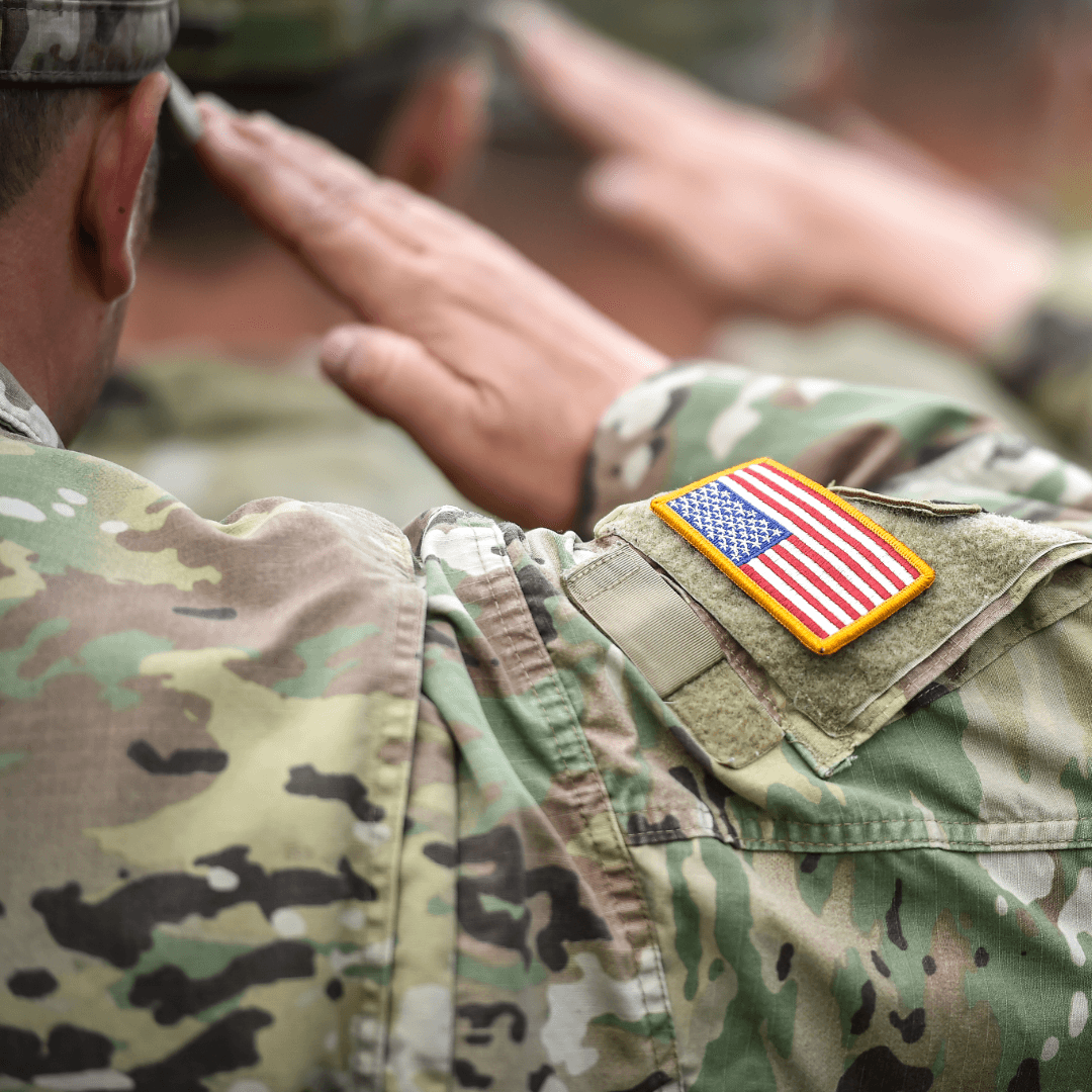 A soldier salutes while wearing a camouflaged uniform with an American flag patch.