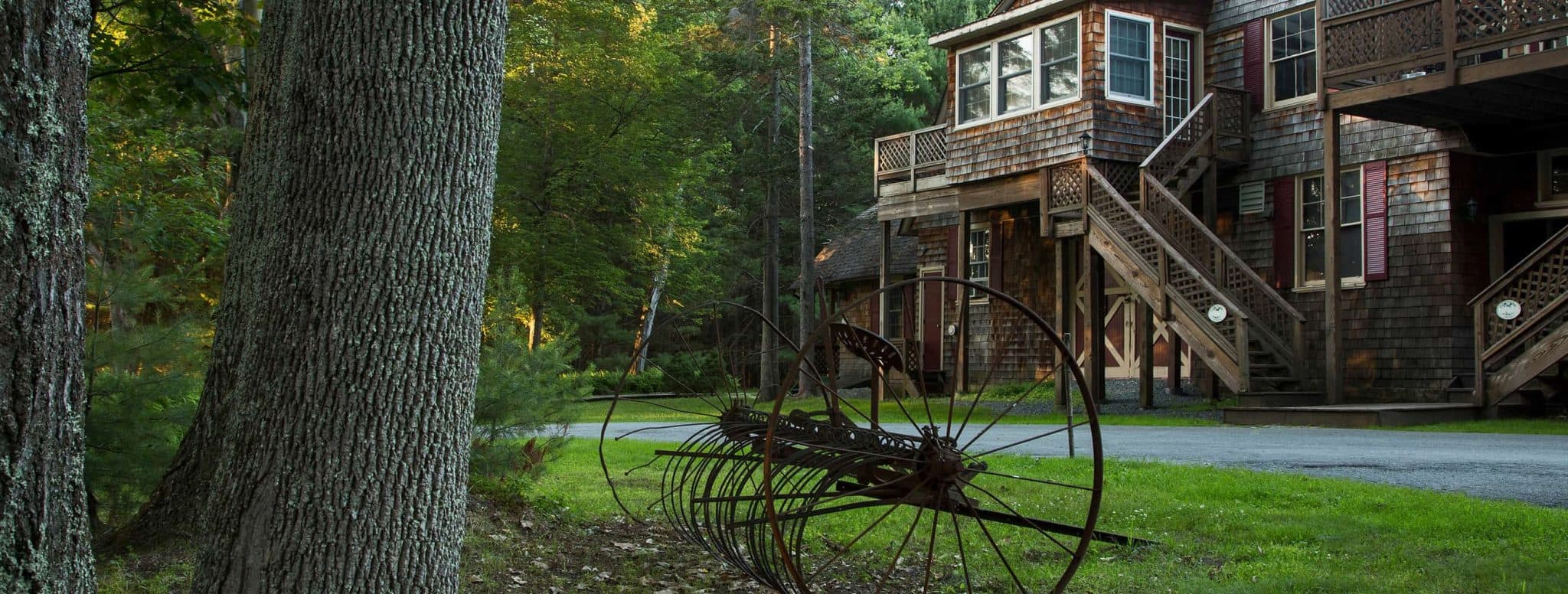 A rusty old farming tool sits in the foreground of a rustic wooden house surrounded by trees.