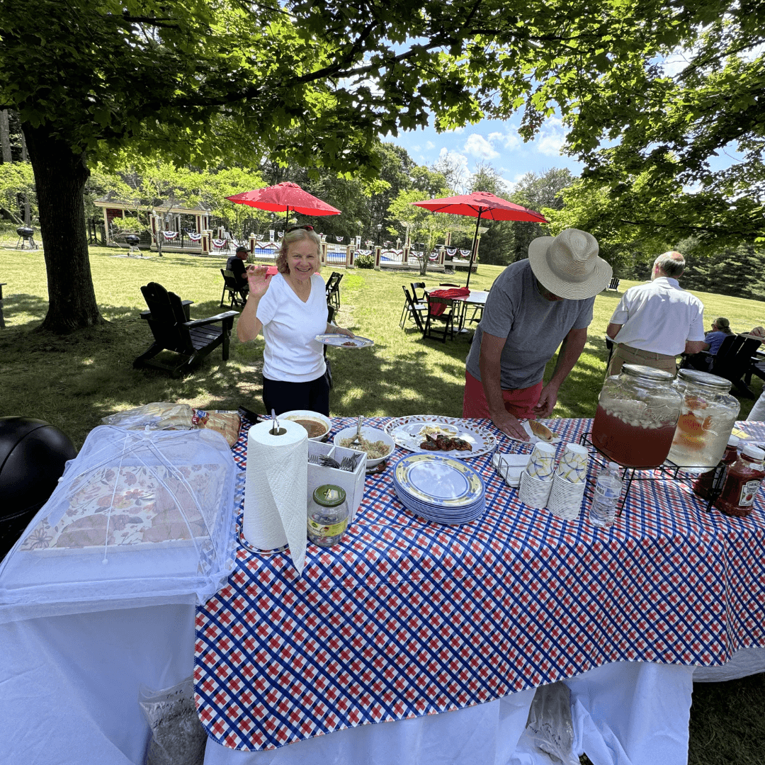 A cheerful woman holds a plate at a picnic table filled with food, while a man prepares dishes in a sunny outdoor setting.