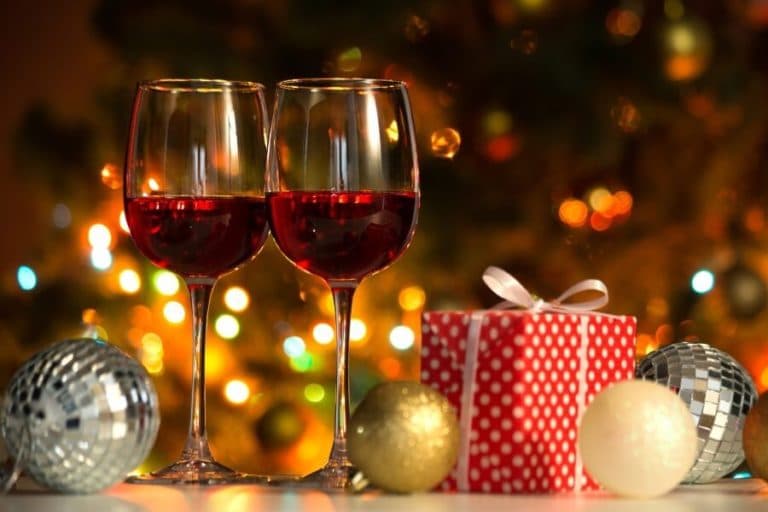 Two glasses of red wine sit beside a festive gift and colorful ornaments, with a softly glowing Christmas tree in the background.