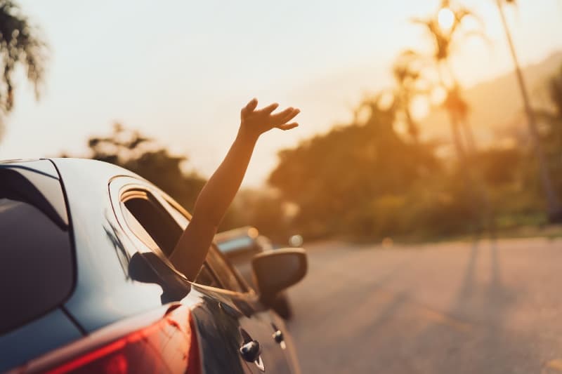 A hand waves out of a car window against a sunset backdrop.