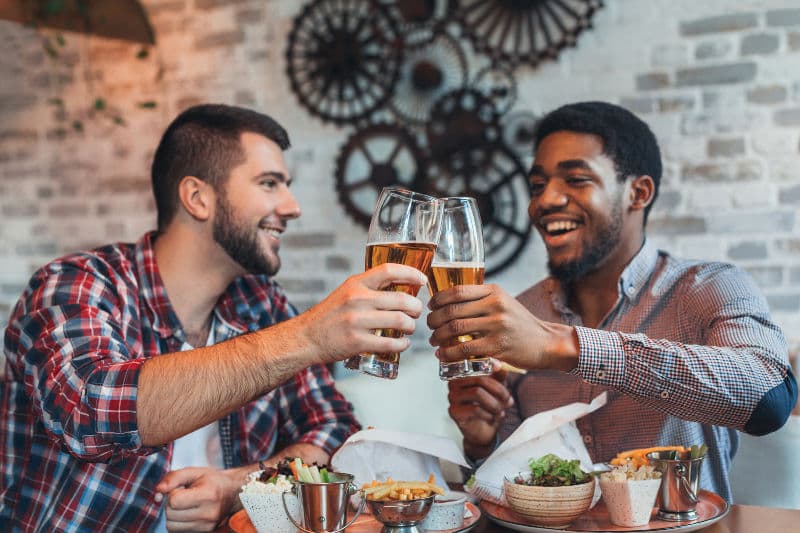Two friends cheers with glasses of beer at a table filled with food.