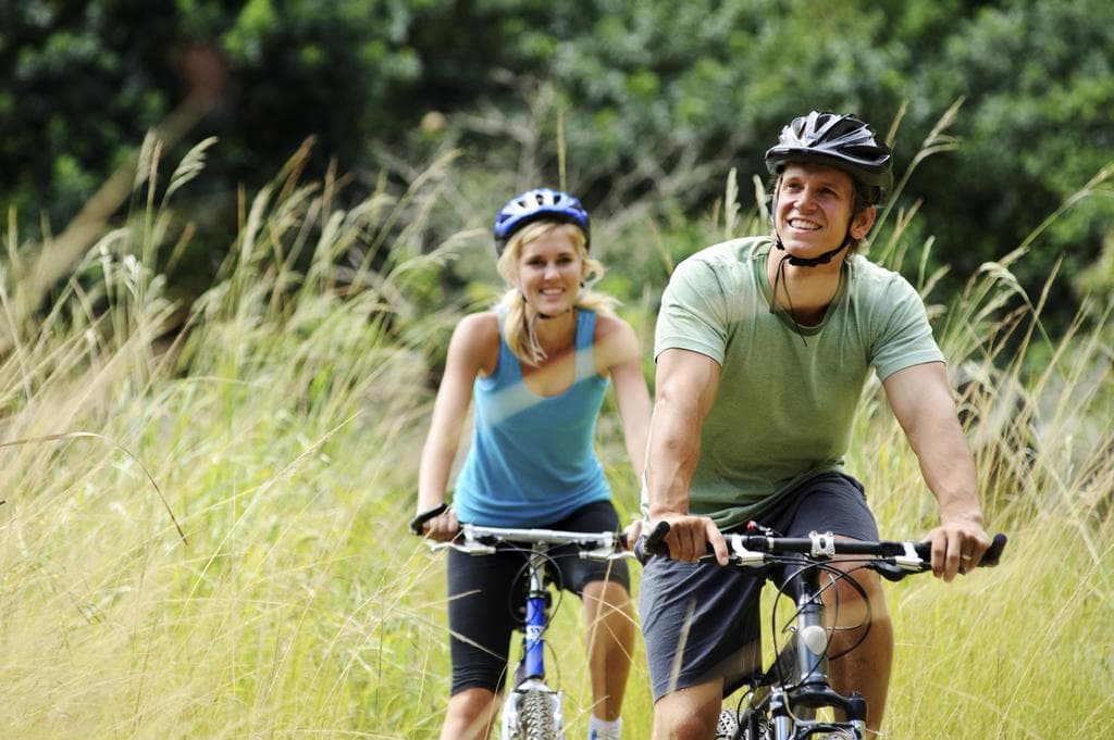A man and a woman smile while riding bicycles through tall grass.